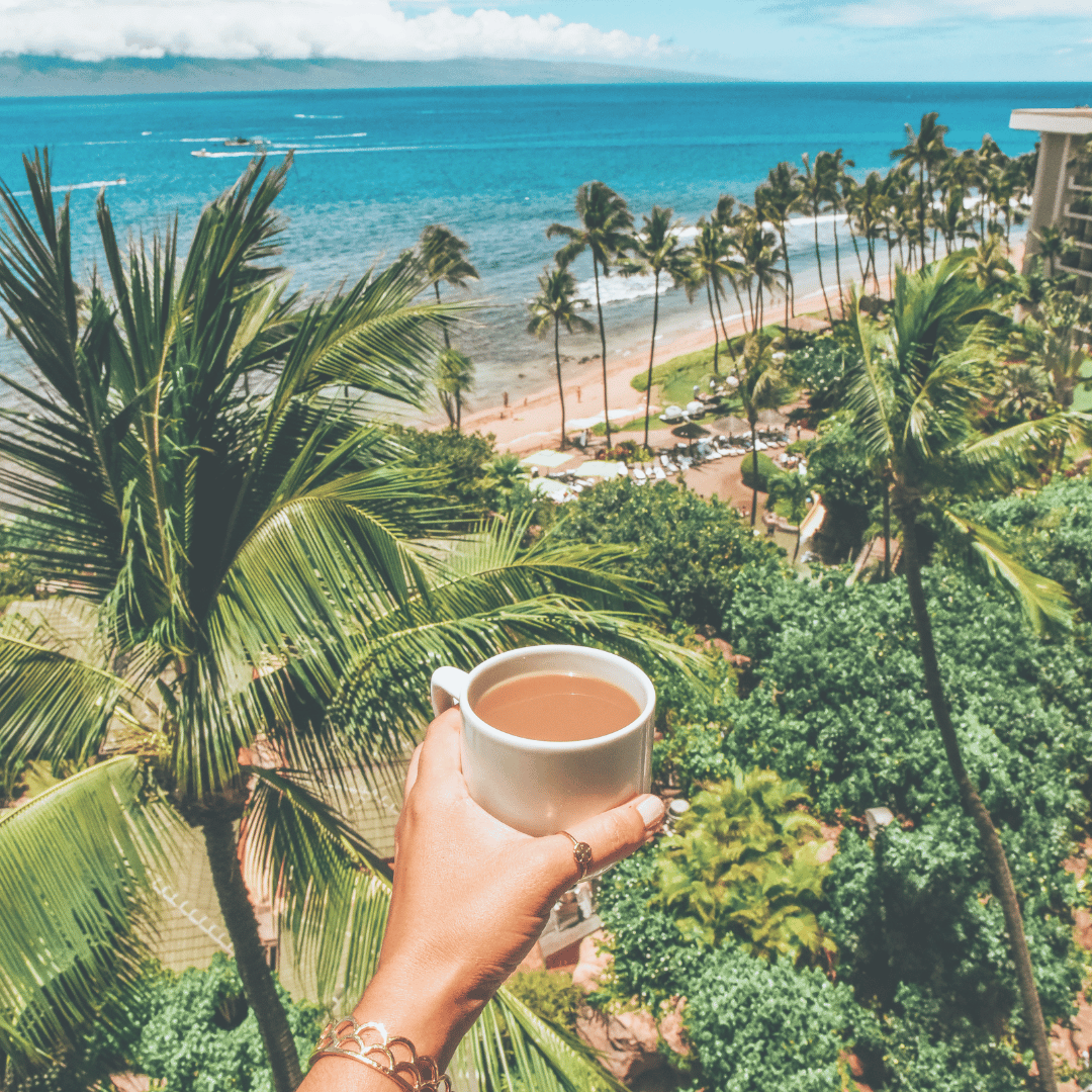 Hand holding a white coffee cup with a tropical beach view in the background, featuring lush green palm trees, blue ocean waters, and a sunny sky.