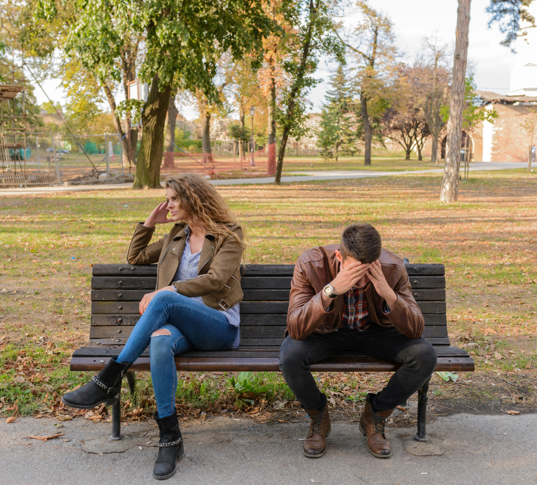 A couple sits on a park bench facing away from each other, both looking upset. The woman has her hand on her head while the man leans forward with his face in his hands, suggesting conflict or emotional tension.